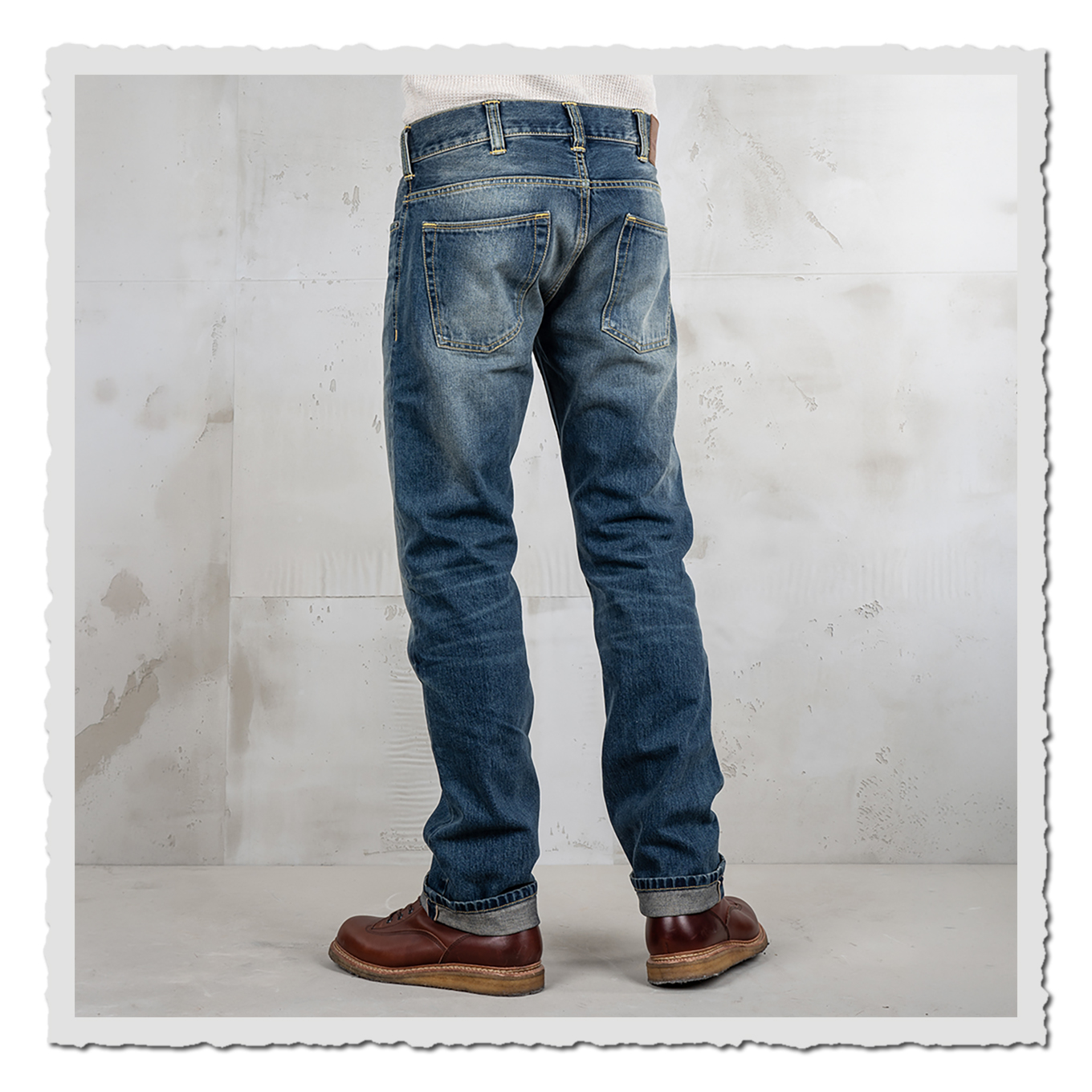 Rückansicht einer getragenen blauen Jeans mit zwei Gesäßtaschen und umgeschlagenen Säumen vor heller Studiowand.
